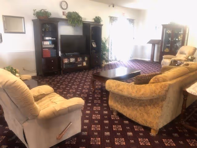 A cozy living room with patterned carpet, a beige recliner, a yellow patterned sofa, a wooden coffee table, and a dark wood entertainment center with a TV and shelves. There are plants on top of the entertainment center and a glass-front cabinet to the right. Sunlight is coming through the windows with sheer curtains.