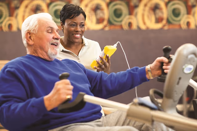 An elderly man using a rowing exercise machine while a smiling female caregiver or trainer holds a yellow device and watches him in a fitness or rehabilitation setting.
