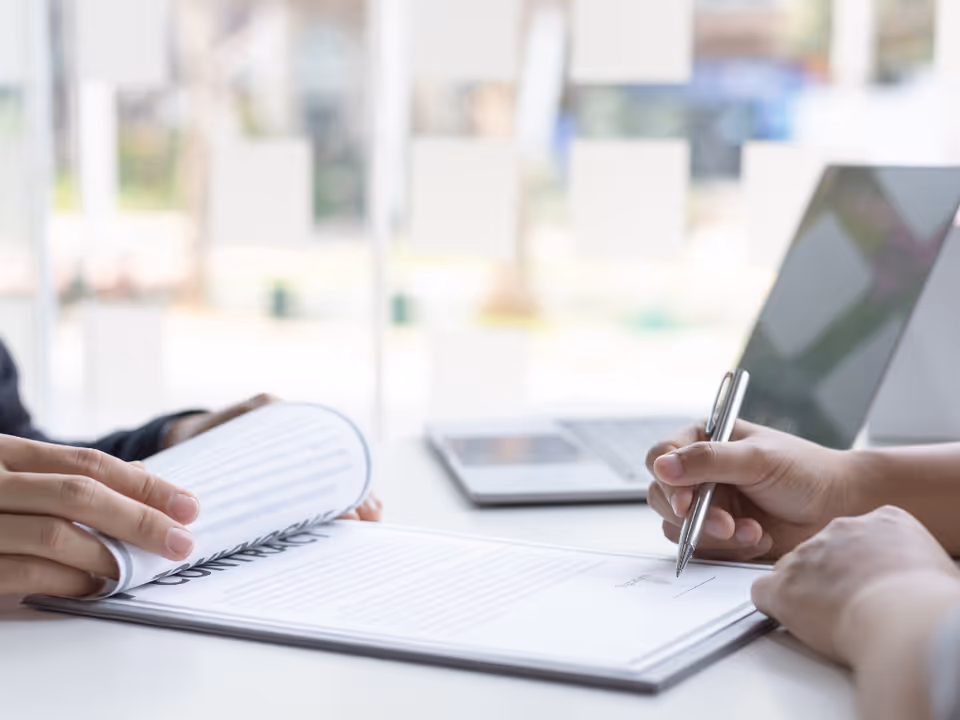 Two people sign documents at a desk with a laptop in an office.