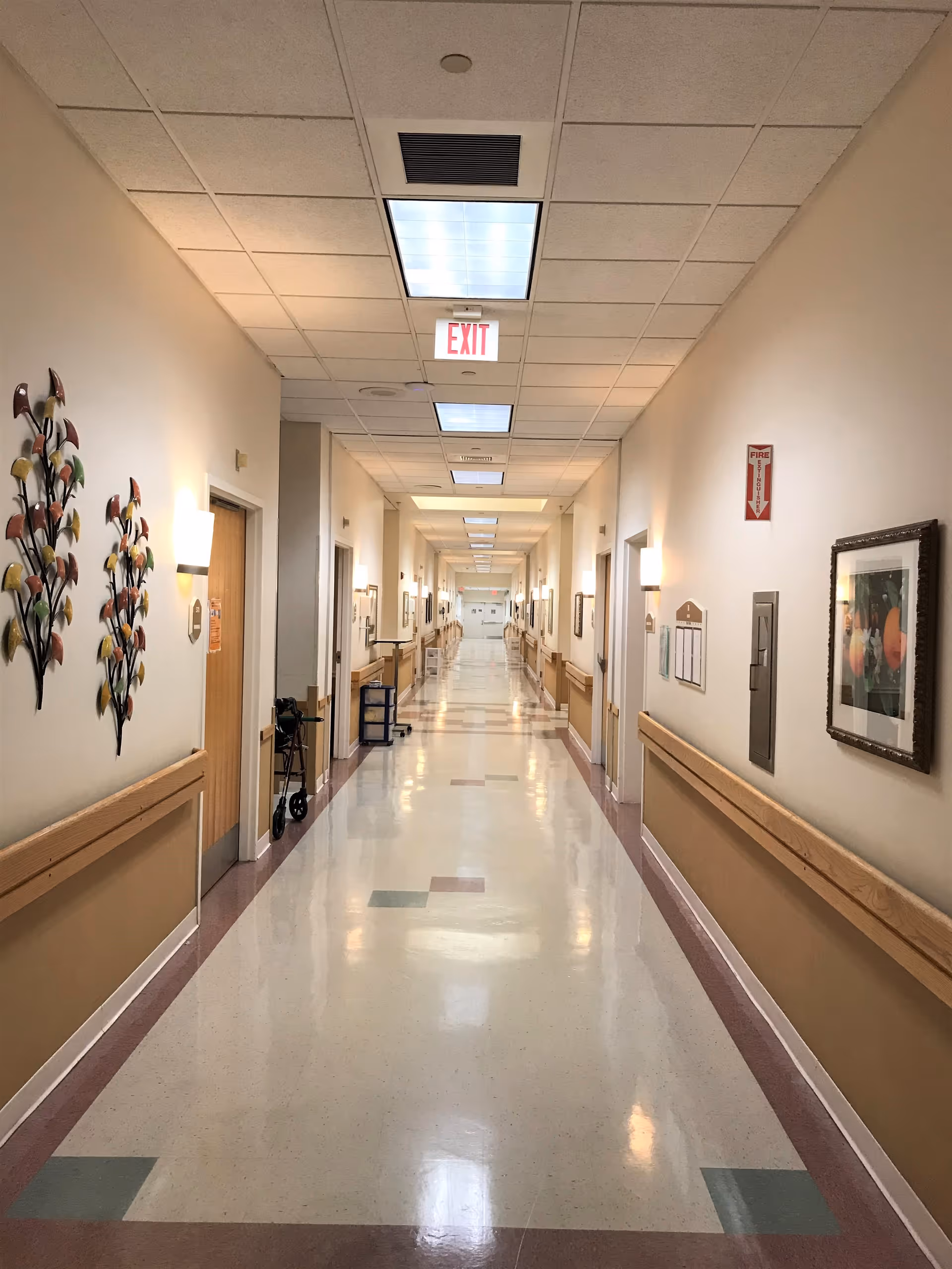A long, clean hallway in a healthcare facility with beige walls, handrails on both sides, wall-mounted lights, decorative wall art, and an exit sign on the ceiling. Doors line both sides of the corridor, and the floor has a polished, tiled surface with colored square accents.