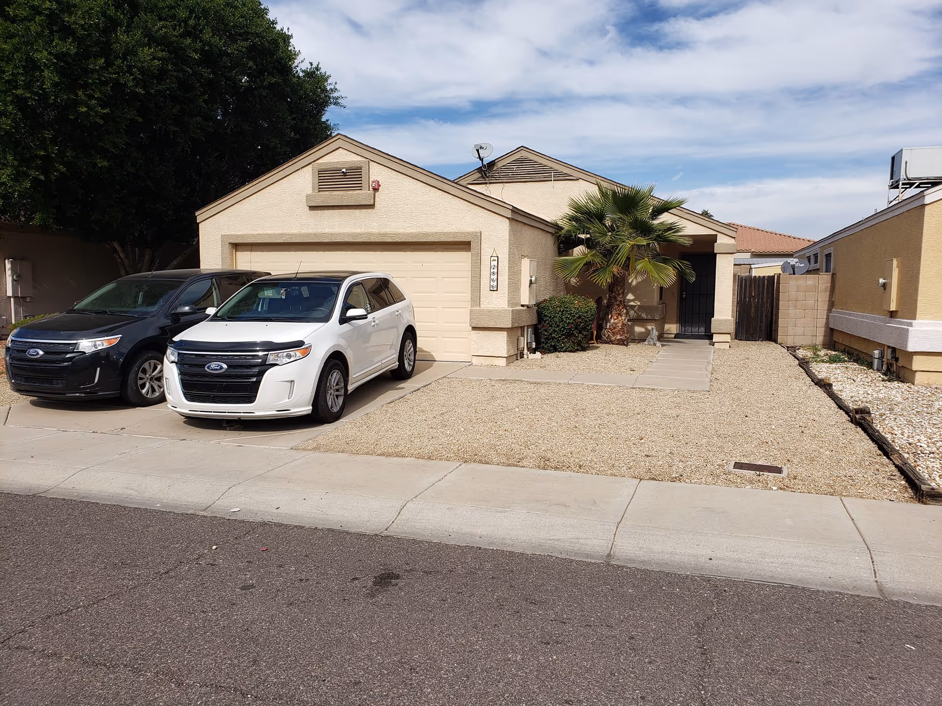 Front exterior of a single-story beige house with a two-car garage, two cars parked in the driveway, and a gravel yard with a small palm tree.