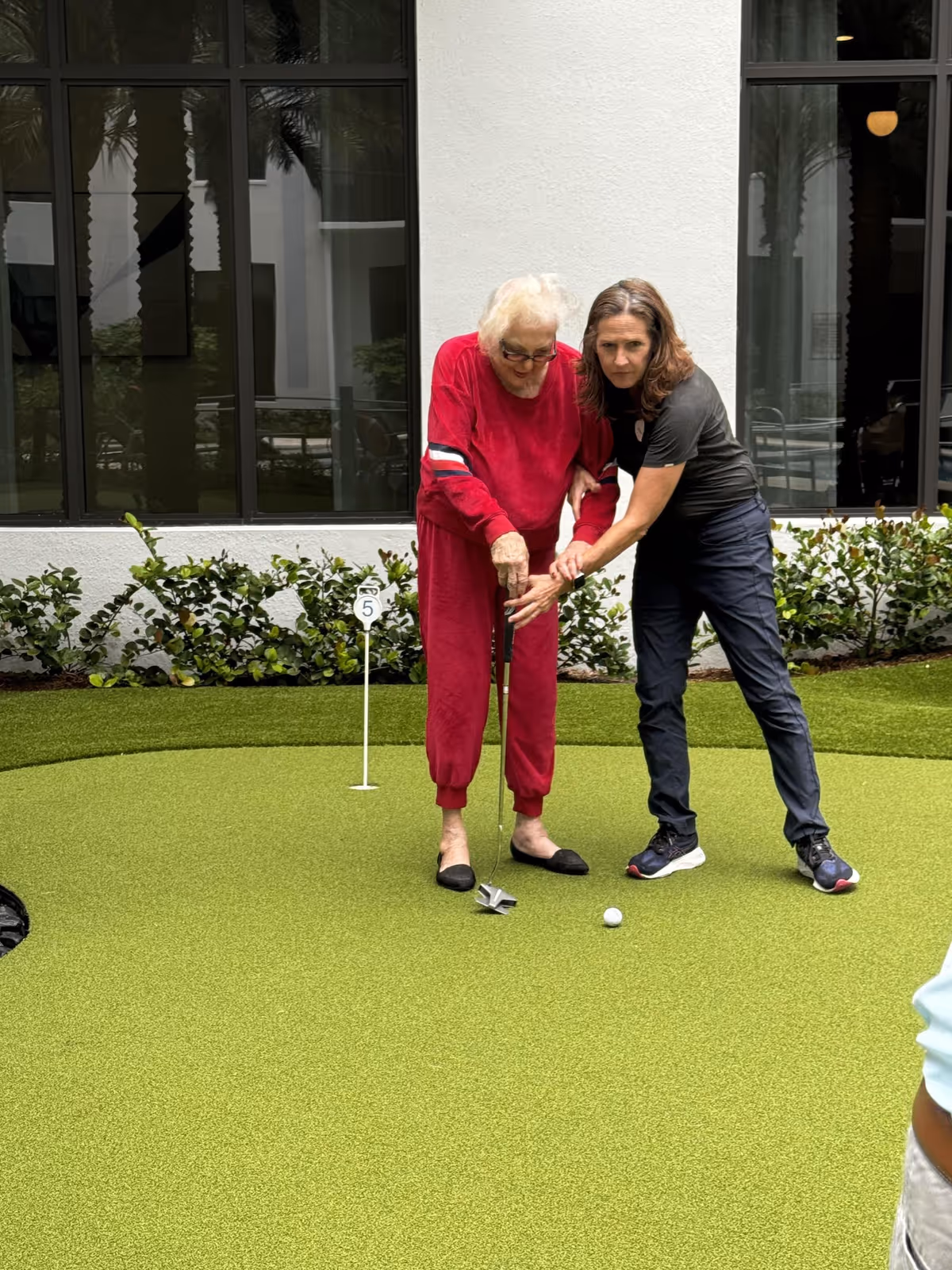 An elderly woman in a red outfit is being assisted by a younger woman as they play mini golf on a putting green outside a building with large windows and greenery.