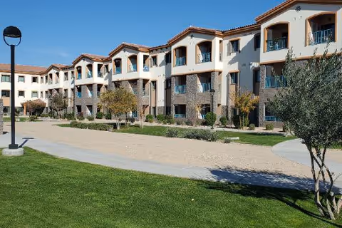 Exterior view of a multi-story senior living facility building with balconies, surrounded by a landscaped area with grass, trees, and a walking path under a clear blue sky.