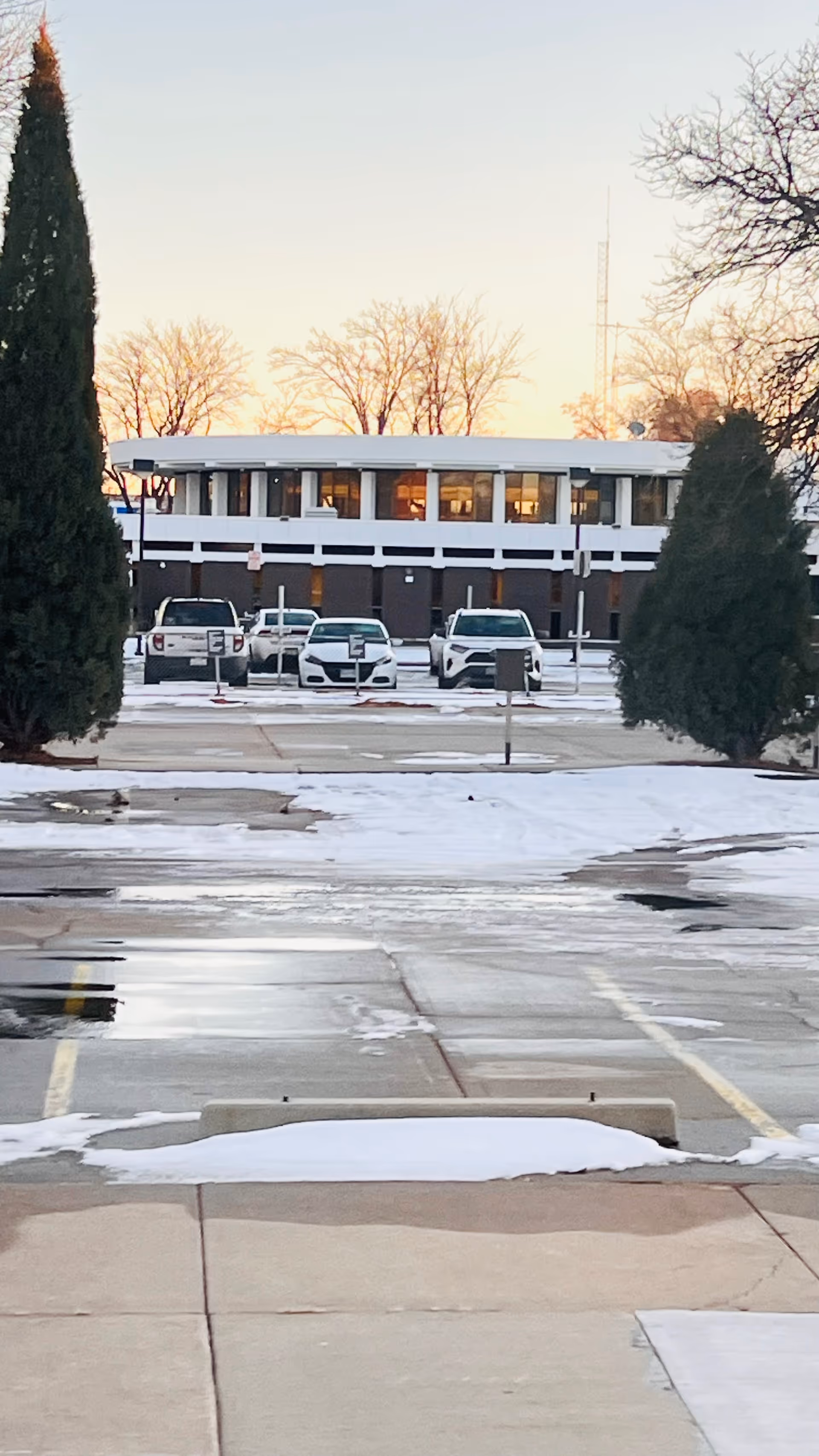 Two tall evergreen trees frame a two-story medical center with a snow-dusted parking lot.