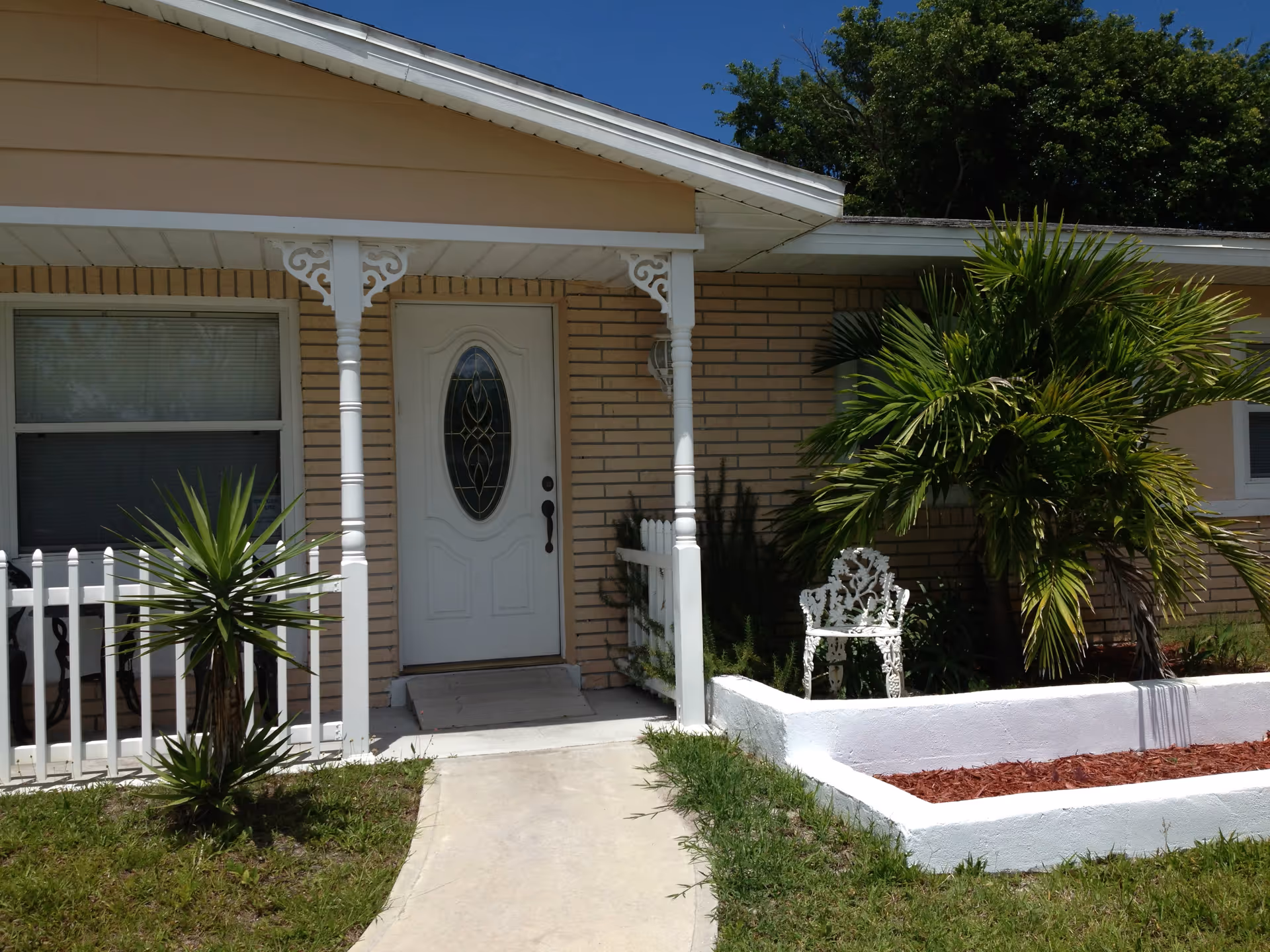 Front exterior of a brick house with a white door, porch pillars, and palm plants.