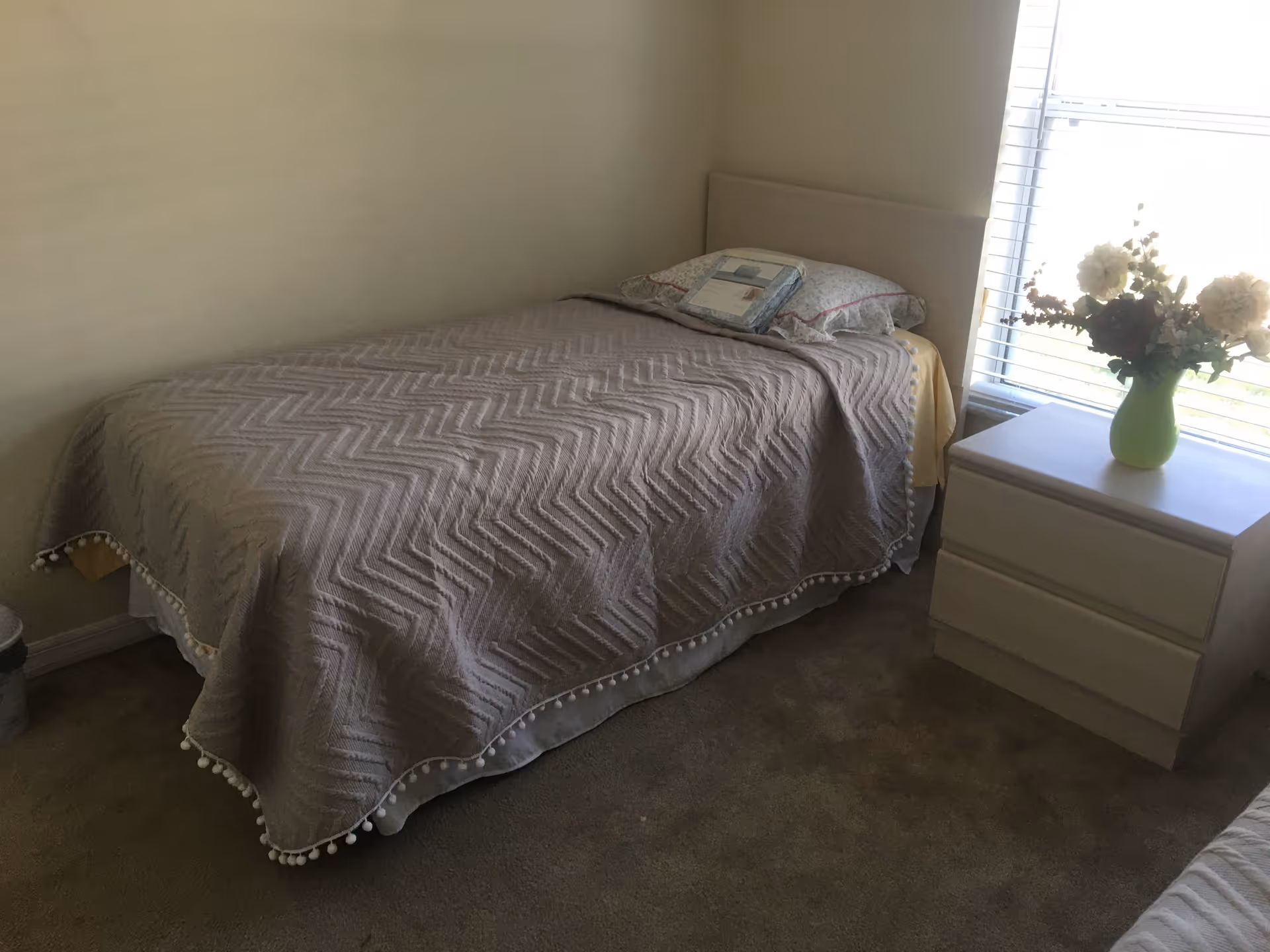 A tidy bedroom with a single bed, nightstand, and vase by a window.
