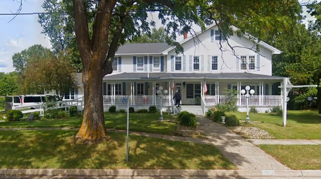 Two-story white building with a wraparound front porch, lawn, large tree and a walkway leading to the entrance.