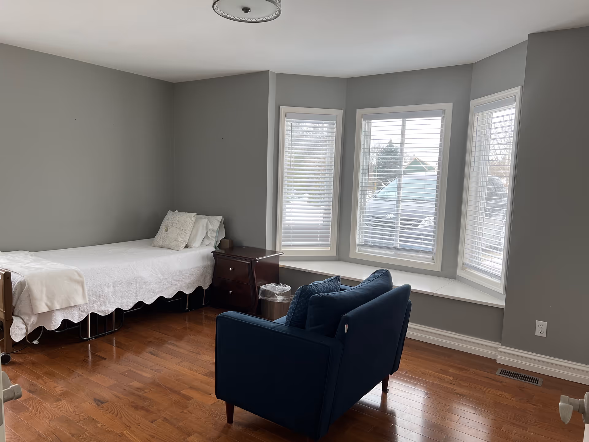A small gray-walled bedroom with a bed, nightstand, and blue armchair by a bay window.