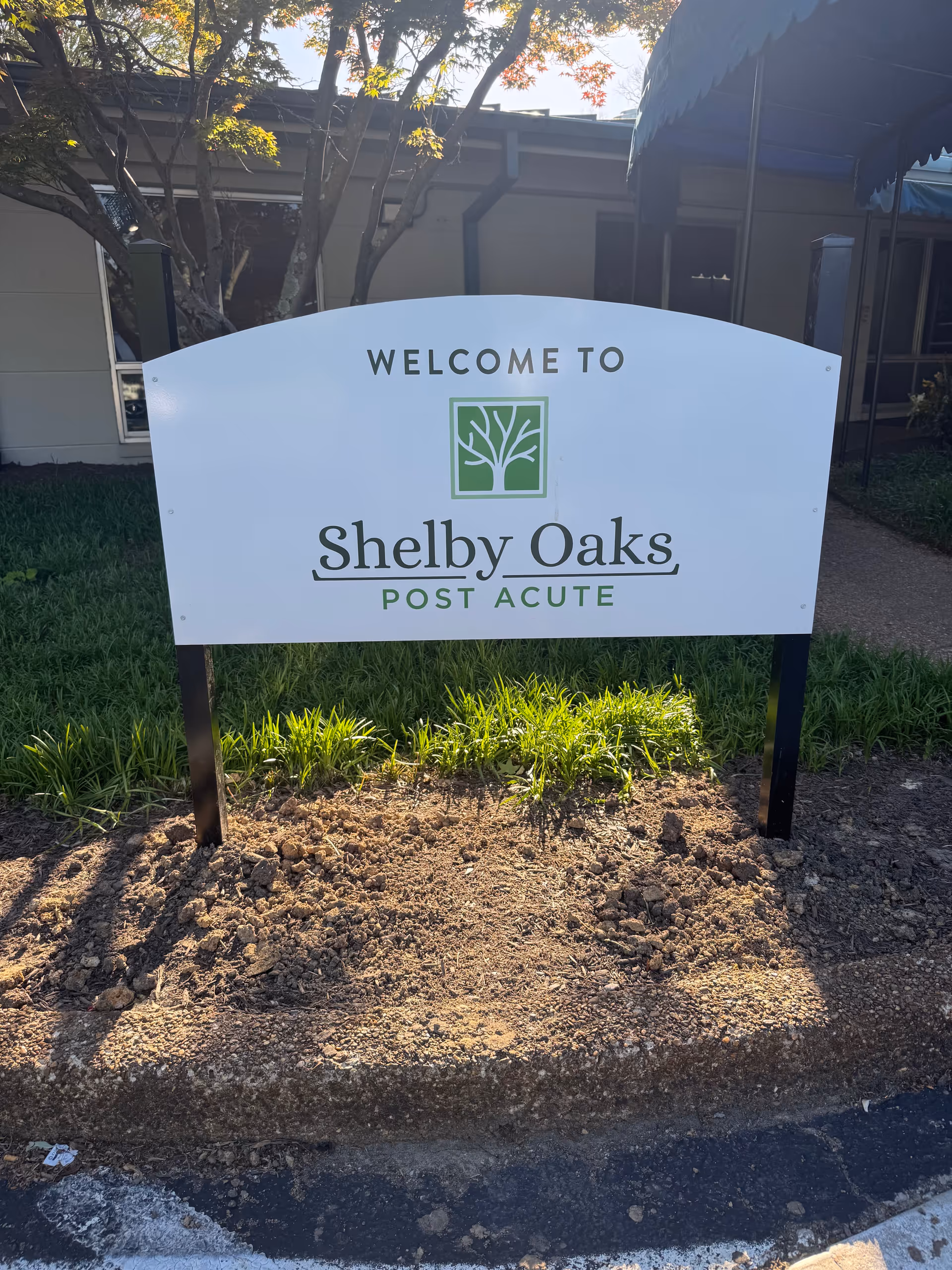 White welcome sign for Shelby Oaks Post Acute stands by a landscaped bed near a building