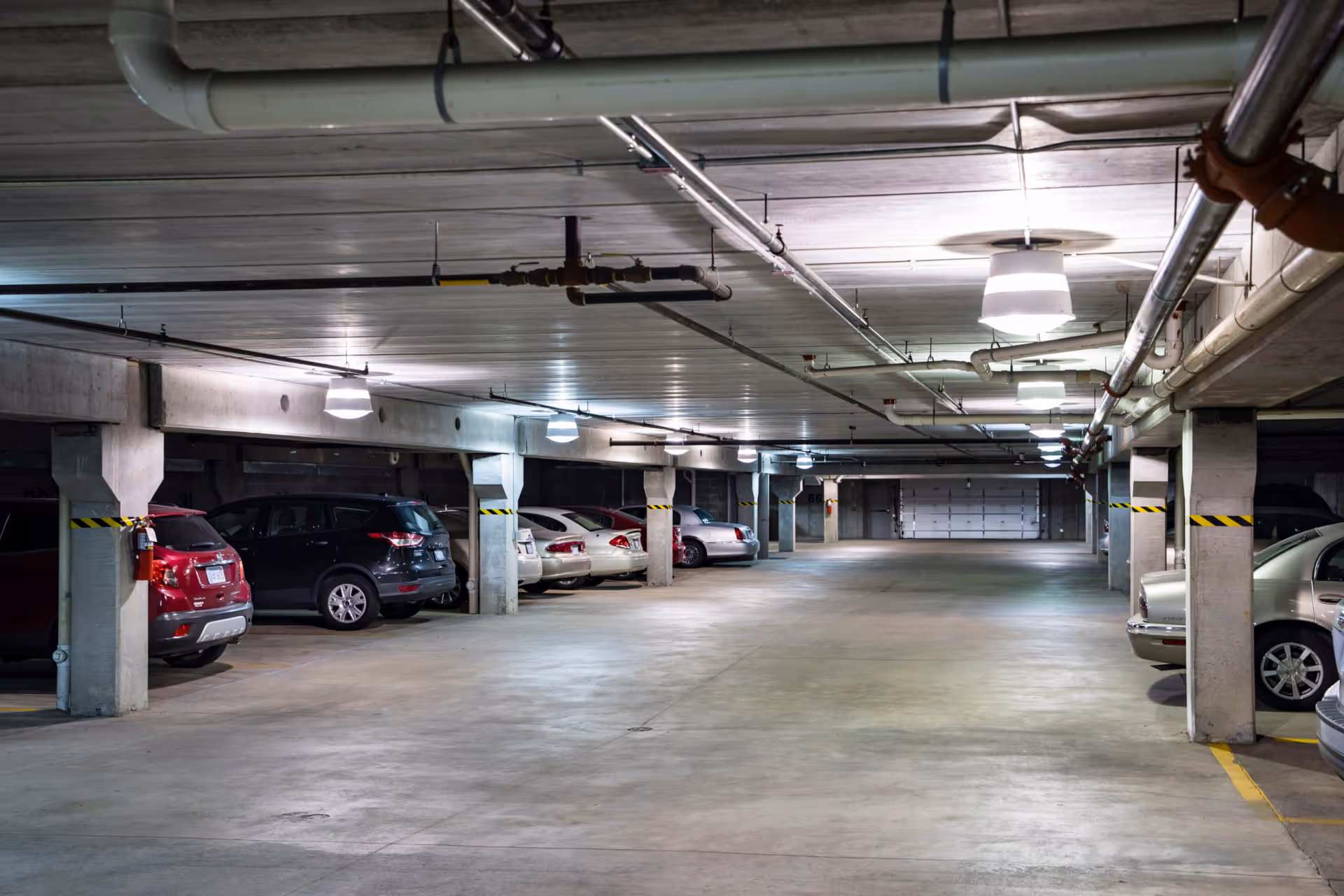 Indoor parking garage with several parked cars along the left and right sides, concrete pillars, and ceiling pipes and lights.