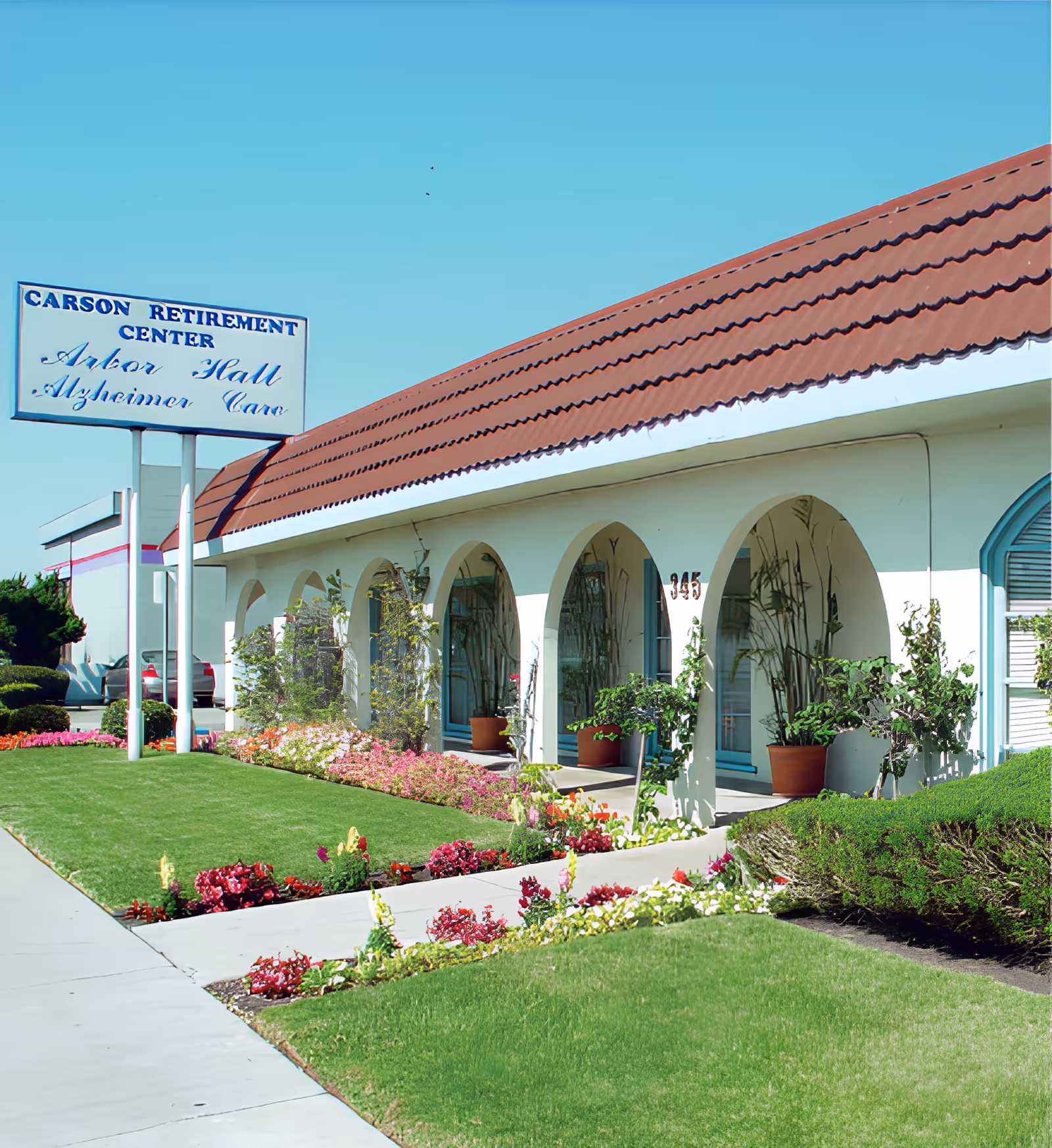 Exterior view of Carson Retirement Center with a red tiled roof and white walls featuring arched doorways. The building is surrounded by well-maintained green lawns and colorful flower beds. A sign near the entrance reads 'Carson Retirement Center Arbor Hall Alzheimer Care'.