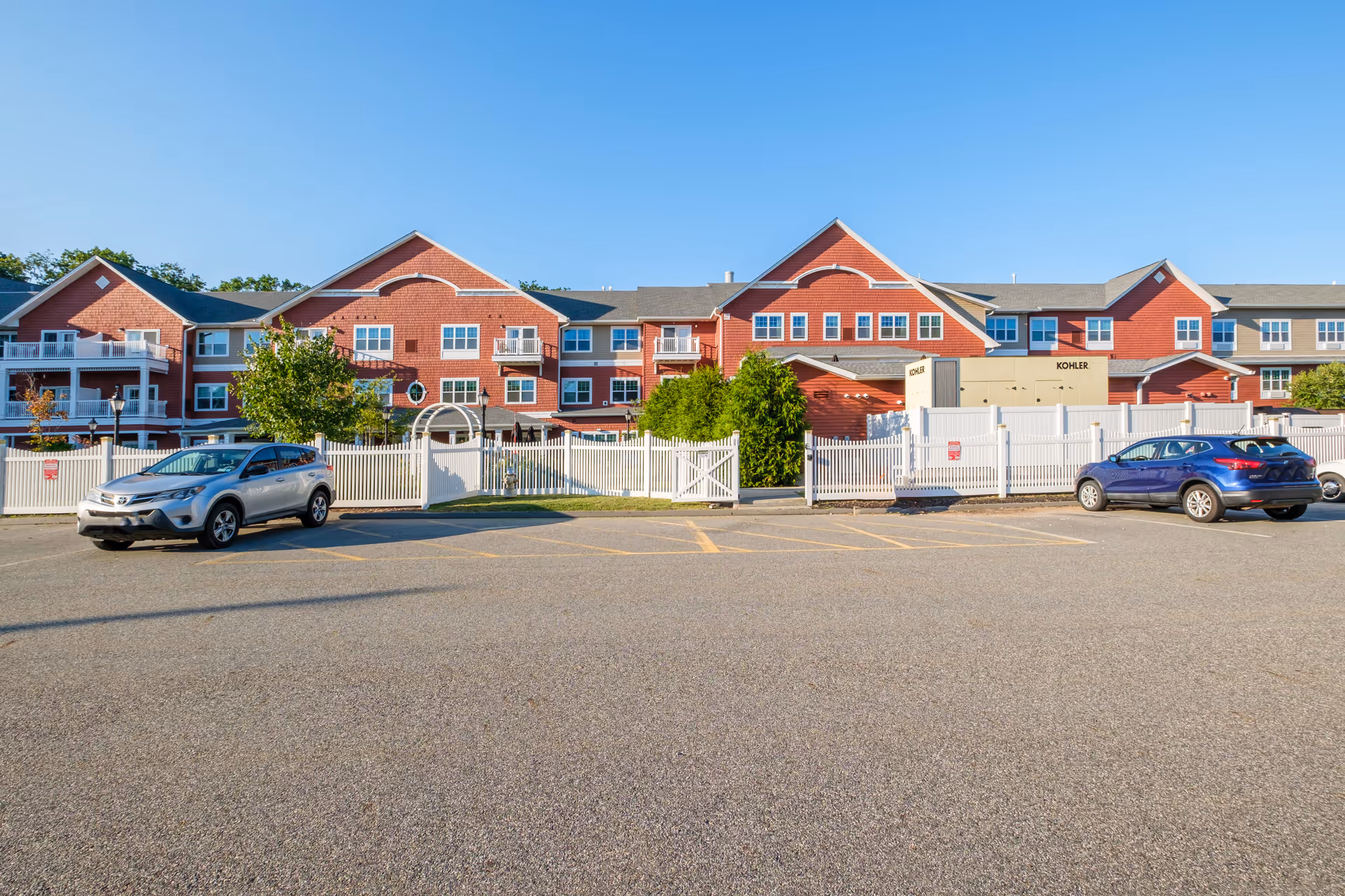 Exterior view of a senior living facility with red brick buildings, white fencing, and a parking lot with two cars parked. The sky is clear and blue.