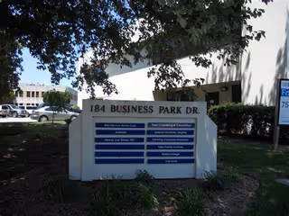 Exterior view of a stone directory sign for 184 Business Park Dr., with surrounding trees.