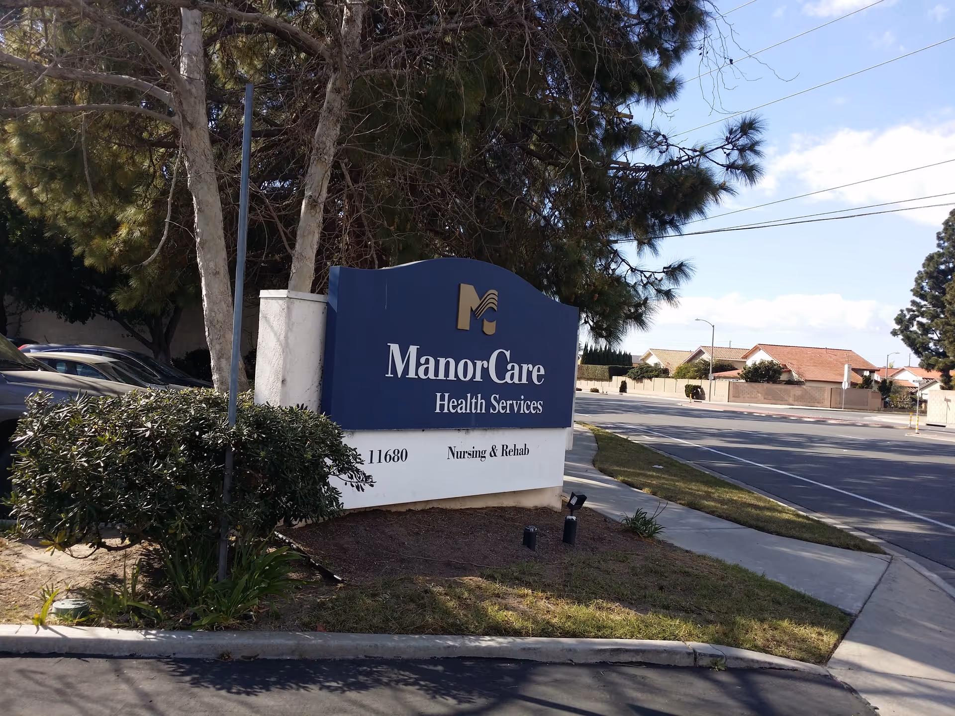 Blue ManorCare Health Services sign at entrance with trees and street in background.