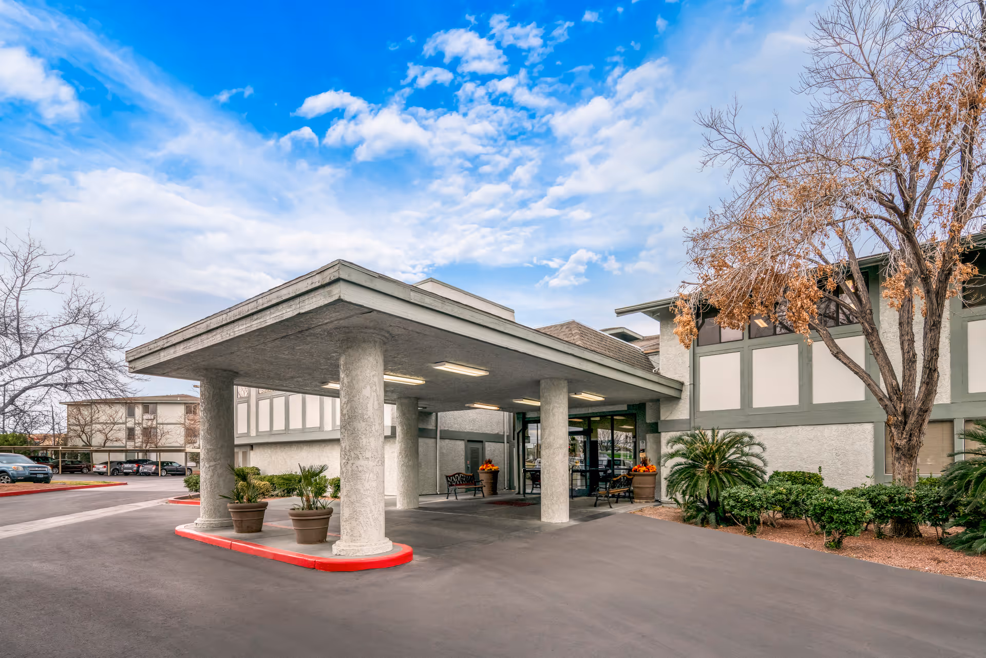Senior living facility entrance with a covered drop-off, columns, and surrounding plants.