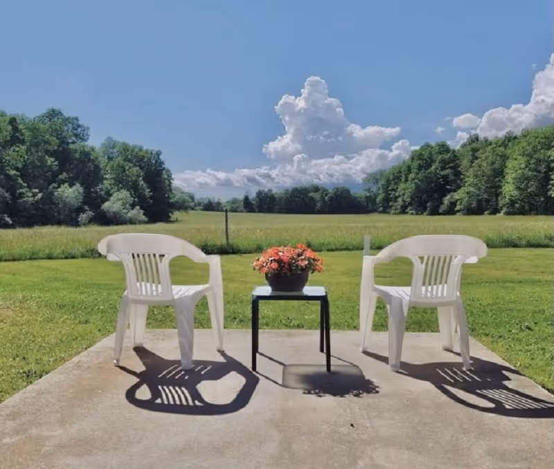 Two white plastic chairs sit on a concrete patio with a table and plant, overlooking fields.