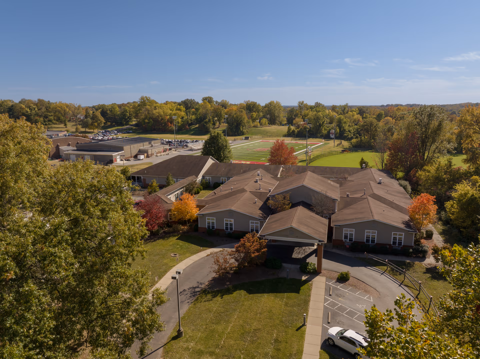Aerial view of a single-story building with a brown roof surrounded by trees with autumn foliage. The building has a covered entrance and a circular driveway with parked cars. In the background, there is a sports field with goalposts and a parking lot. The sky is clear with a few clouds.