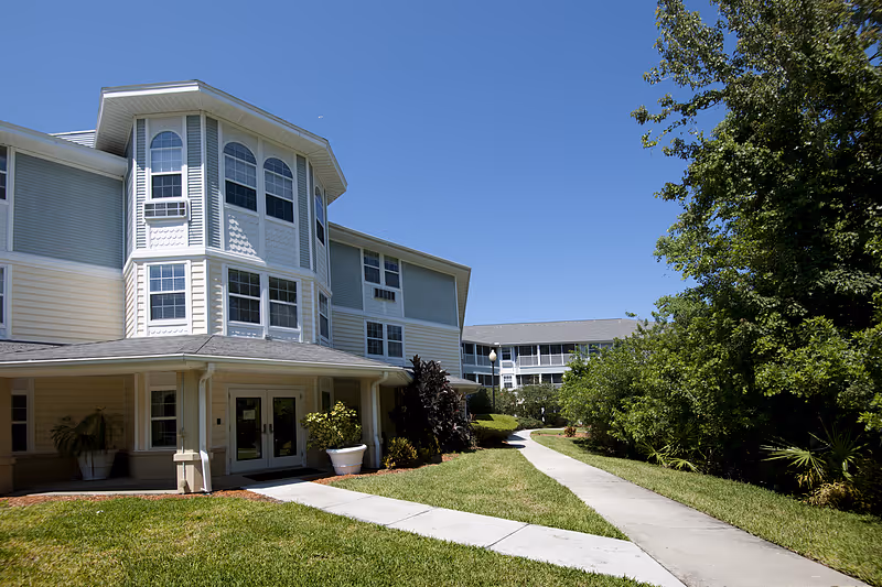 Exterior view of a senior living facility building with light-colored siding and multiple windows under a clear blue sky. A paved walkway curves through a well-maintained lawn and landscaping with trees and shrubs.