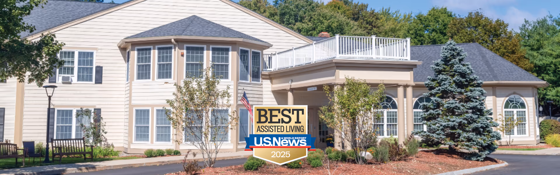 Exterior view of a two-story senior living facility building with beige siding, multiple windows, and a covered entrance. There are trees, shrubs, and benches in the landscaped area in front of the building. A sign in front displays 'Best Assisted Living U.S. News 2025'.
