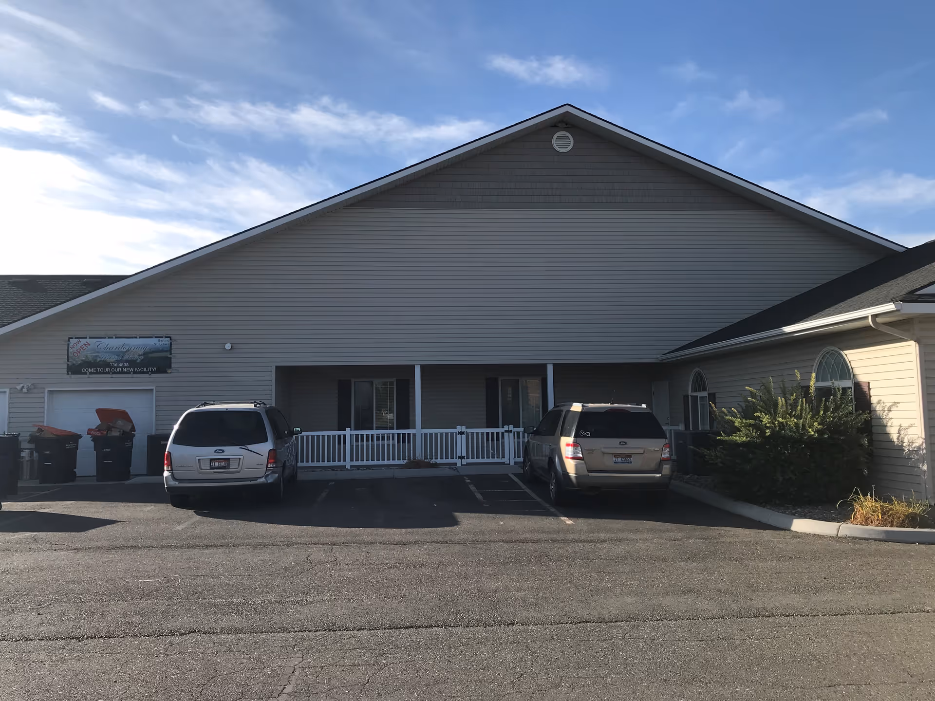 Exterior view of a single-story building with beige siding and a triangular roof. Two vehicles are parked in front of the building, which has a small white fence and two windows with dark shutters. There is a sign on the left side of the building that reads 'Chardonnay Assisted Living - Twin Falls' with additional text about touring the new facility. The sky is partly cloudy.