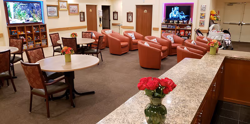Communal lounge and dining area with round tables, upholstered chairs and armchairs arranged around a TV and flowers on the tables and countertop.