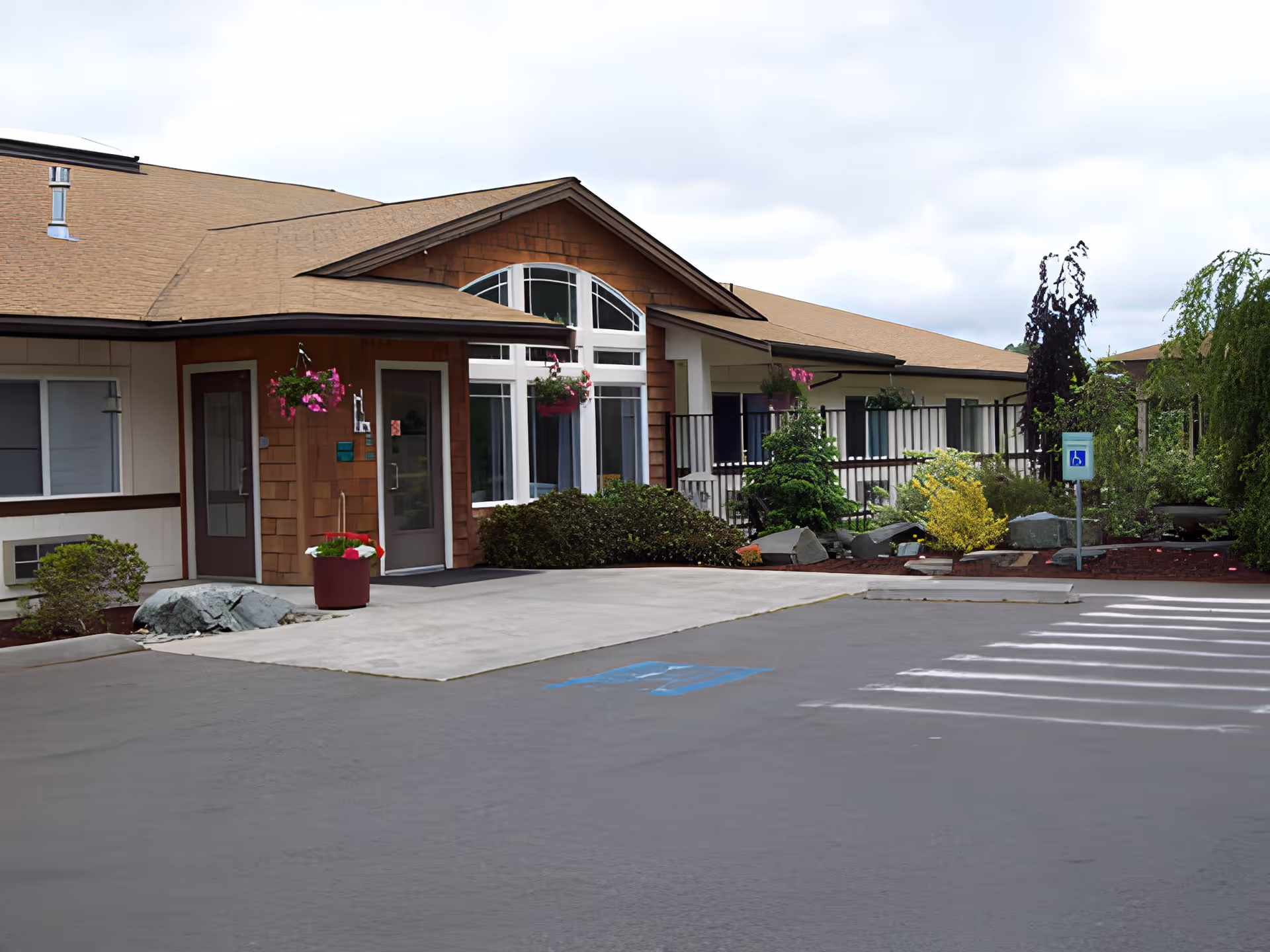 Front entrance of a single-story senior living facility with a covered doorway, hanging flower baskets, landscaped beds, and a handicapped parking space.