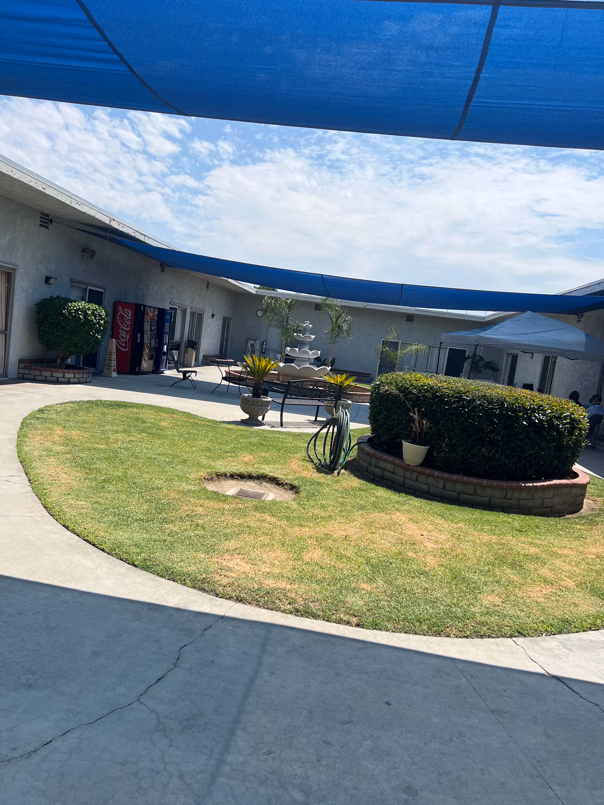Outdoor courtyard with circular grassy area, fountain, seating, and blue shade sails.