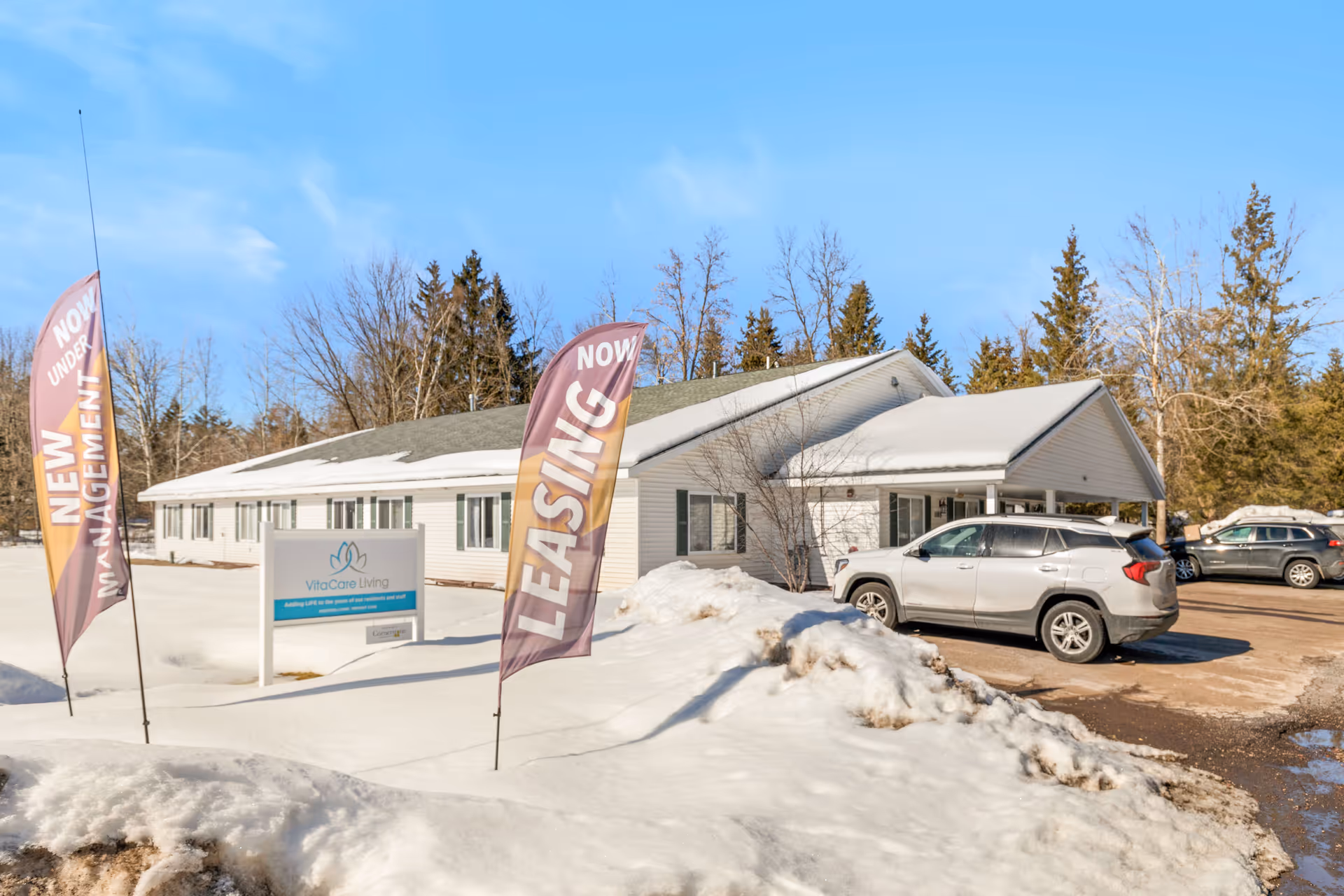 Snow-covered parking lot in front of VitaCare Living Ladysmith facility with leasing flags.