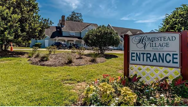 Exterior view of Homestead Village of Pensacola showing a large building with a sloped roof surrounded by green grass, trees, and shrubs under a blue sky. A sign in the foreground reads 'Homestead Village Florida, LLC ENTRANCE'.
