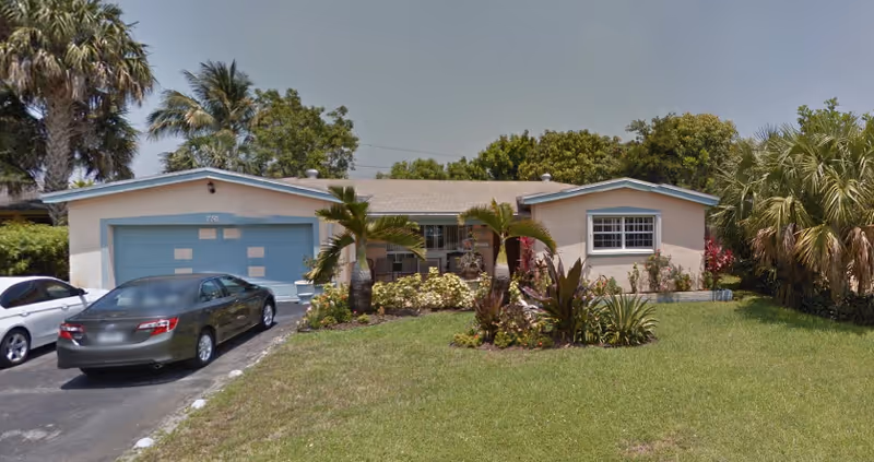 Single-story pastel home with blue garage doors, palm trees, and a driveway.