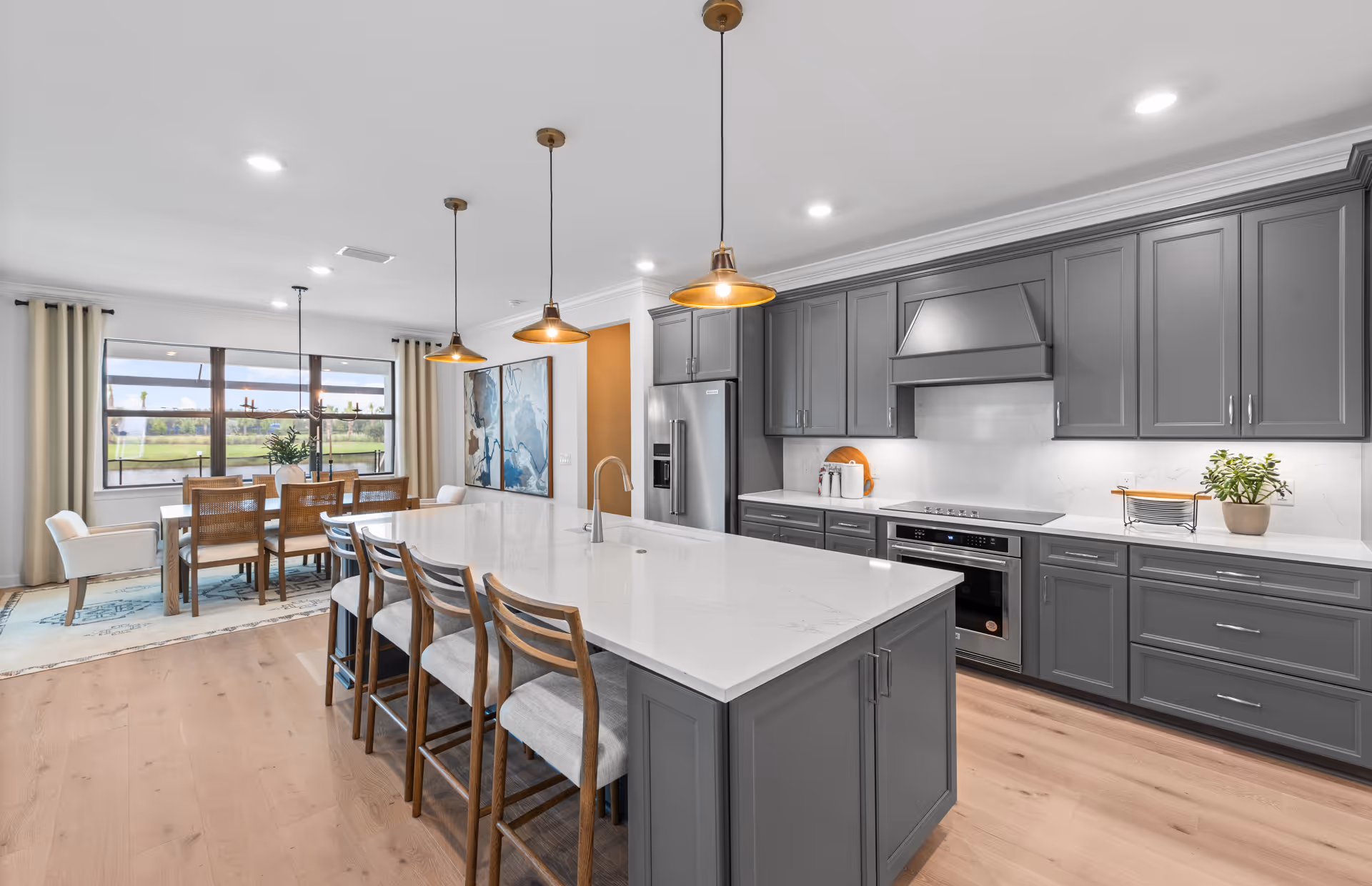 Modern kitchen and dining area in a retirement community home featuring a large white island with four wooden chairs, gray cabinetry, stainless steel appliances, three pendant lights above the island, and a dining table with six chairs near a large window with beige curtains.