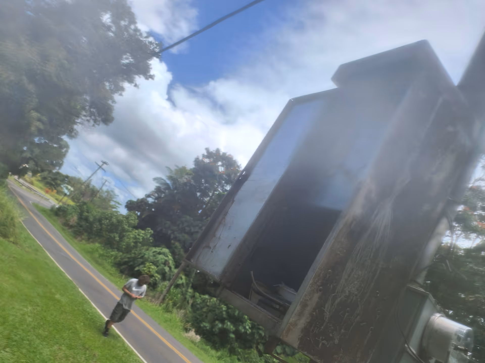A large, rusty metal container on a trailer beside a rural road while a person walks.