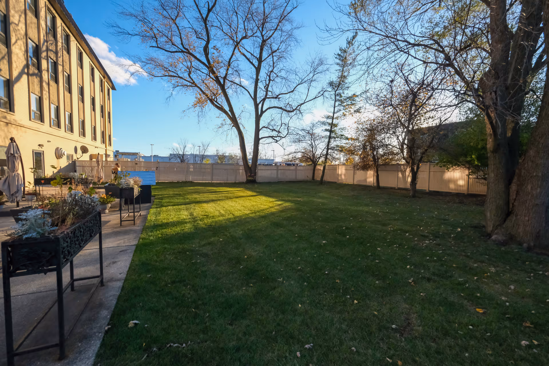Outdoor fenced lawn and patio next to a multi-story senior living building with trees and planters.