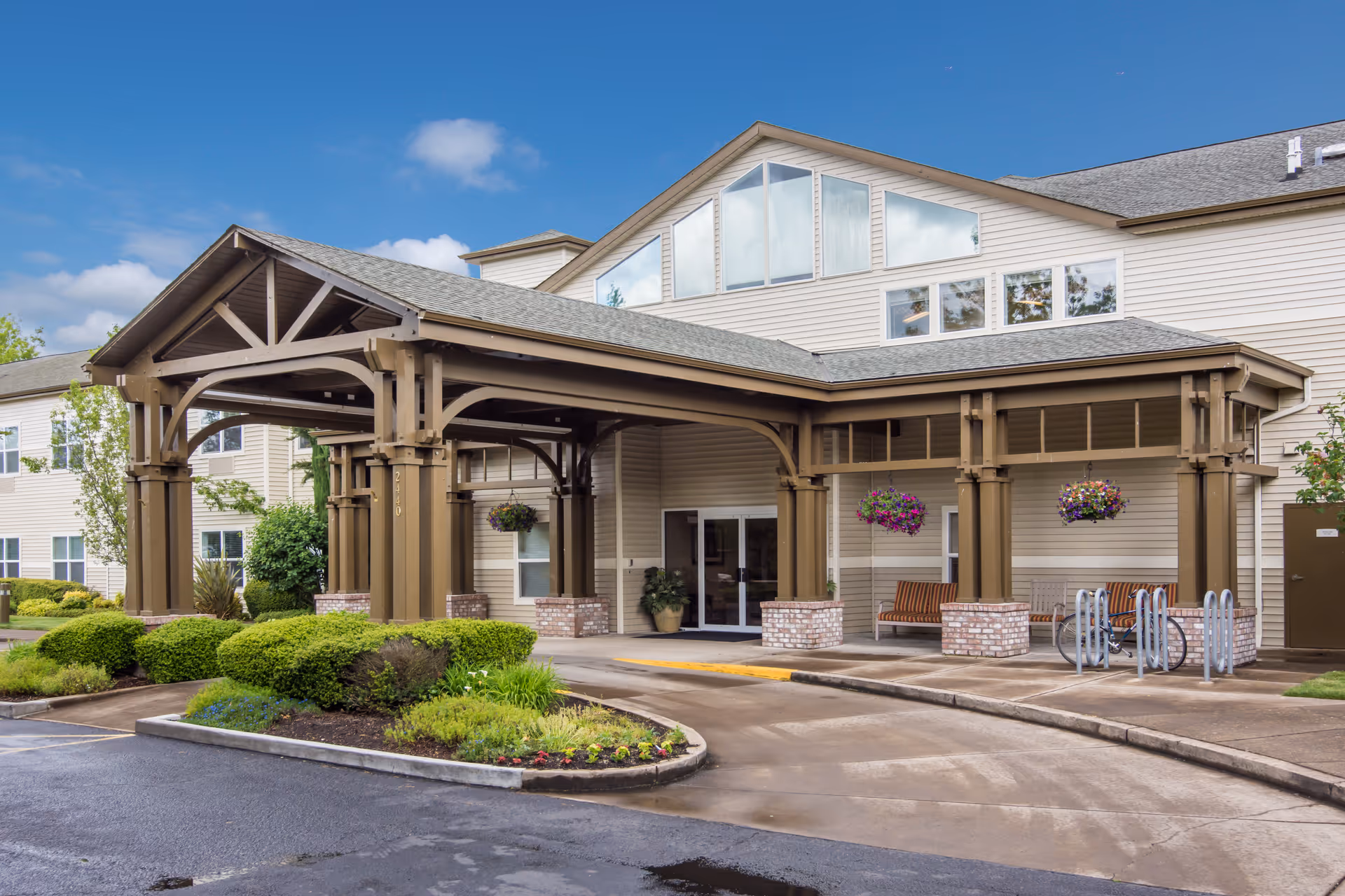 Exterior view of Merrill Gardens at Sheldon Park showing the entrance with a covered driveway supported by large wooden beams. There are hanging flower baskets, benches, a bike rack, and well-maintained landscaping with bushes and flowers. The building has beige siding and multiple windows under a partly cloudy blue sky.