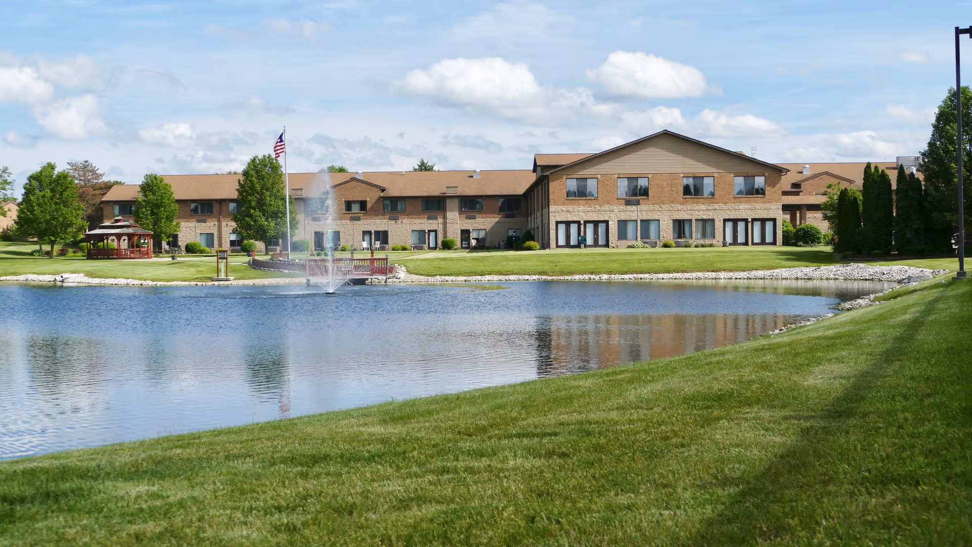 Two-story brick senior living building beside a pond with a fountain, gazebo, flagpole, and a manicured lawn under a blue sky.