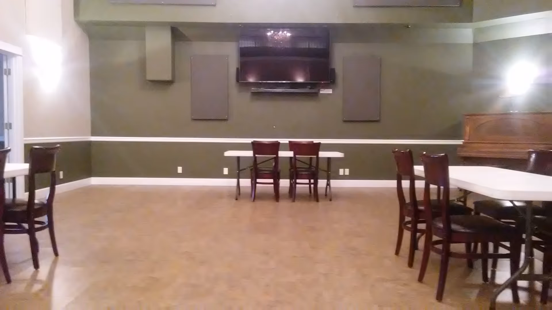 A spacious senior living dining area with tables, chairs, a wall-mounted TV, and a piano