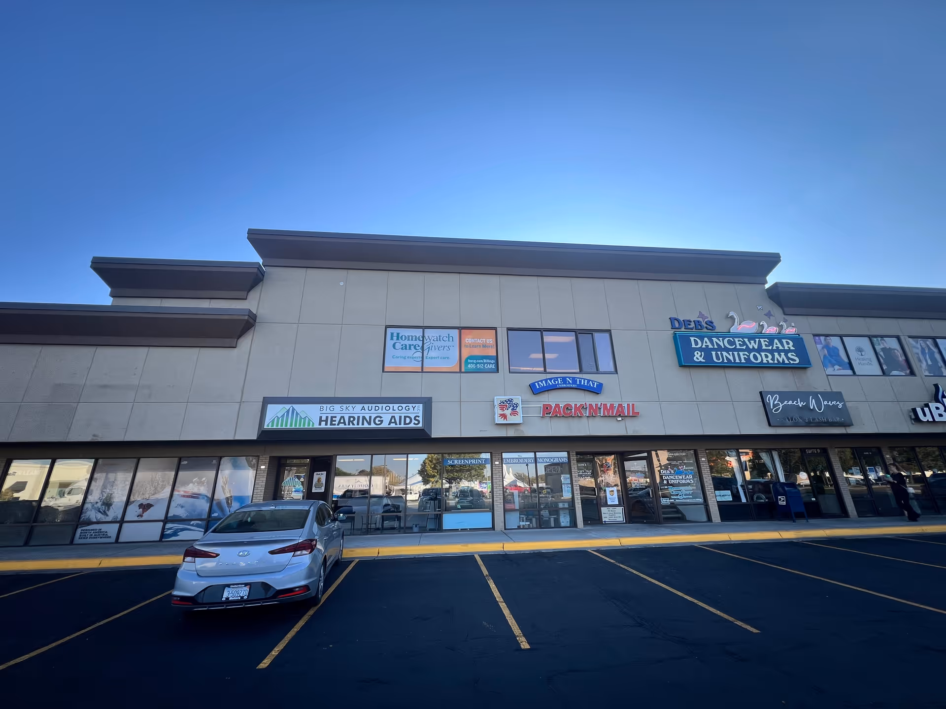Strip mall front with multiple stores and Homewatch CareGivers sign under a blue sky