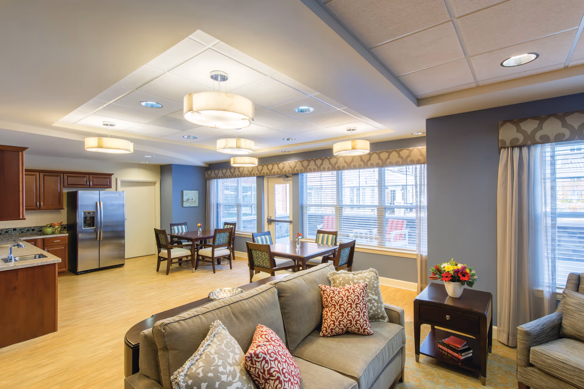 Open-plan senior living common area with sofa, dining tables, and a kitchen.