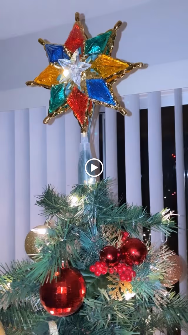 Close-up view of the top of a decorated Christmas tree with a colorful star tree topper featuring red, blue, green, and yellow sections. The tree is adorned with red and gold ornaments and red berries, set against a background of vertical white blinds.