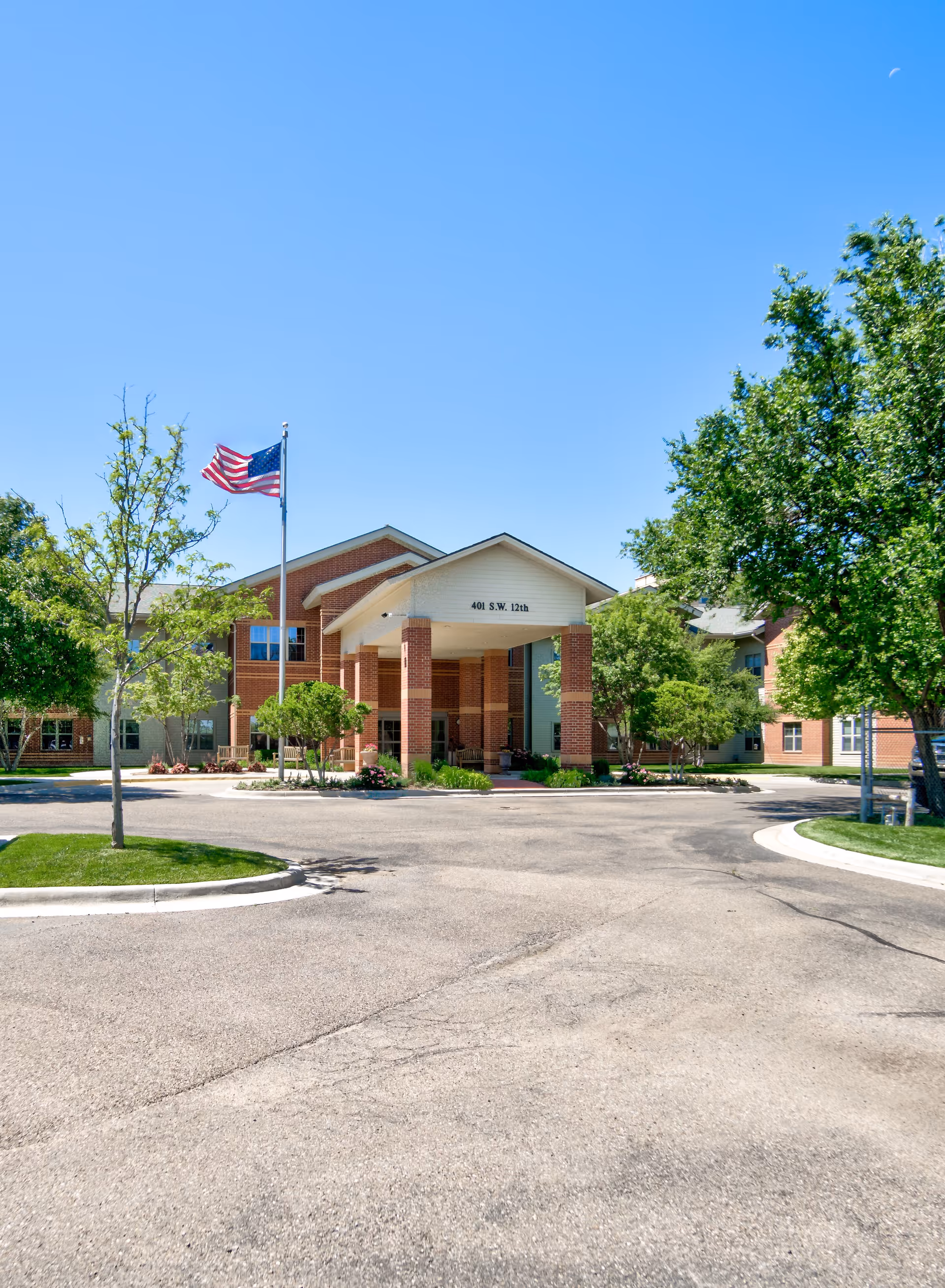 Front exterior view of a senior living facility building with a covered entrance supported by brick columns. An American flag is flying on a flagpole to the left of the entrance. The building is surrounded by trees and landscaping under a clear blue sky.