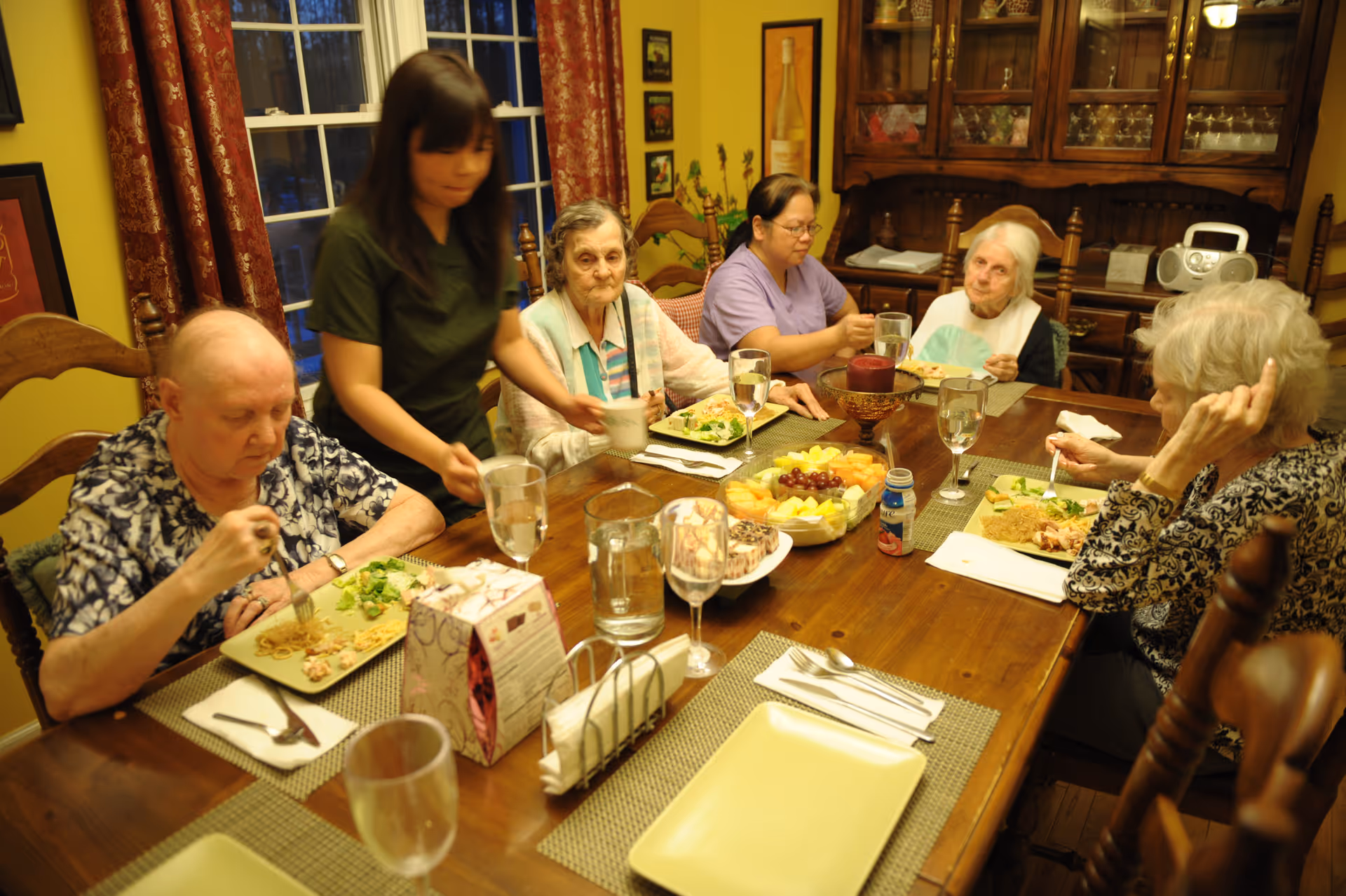 A group of elderly women seated around a wooden dining table having a meal together in a warmly lit room. A caregiver is serving food to one of the women. The table is set with plates of food, glasses of water, a fruit platter, and a tissue box. The room has yellow walls, red patterned curtains, and a wooden cabinet with glassware in the background.