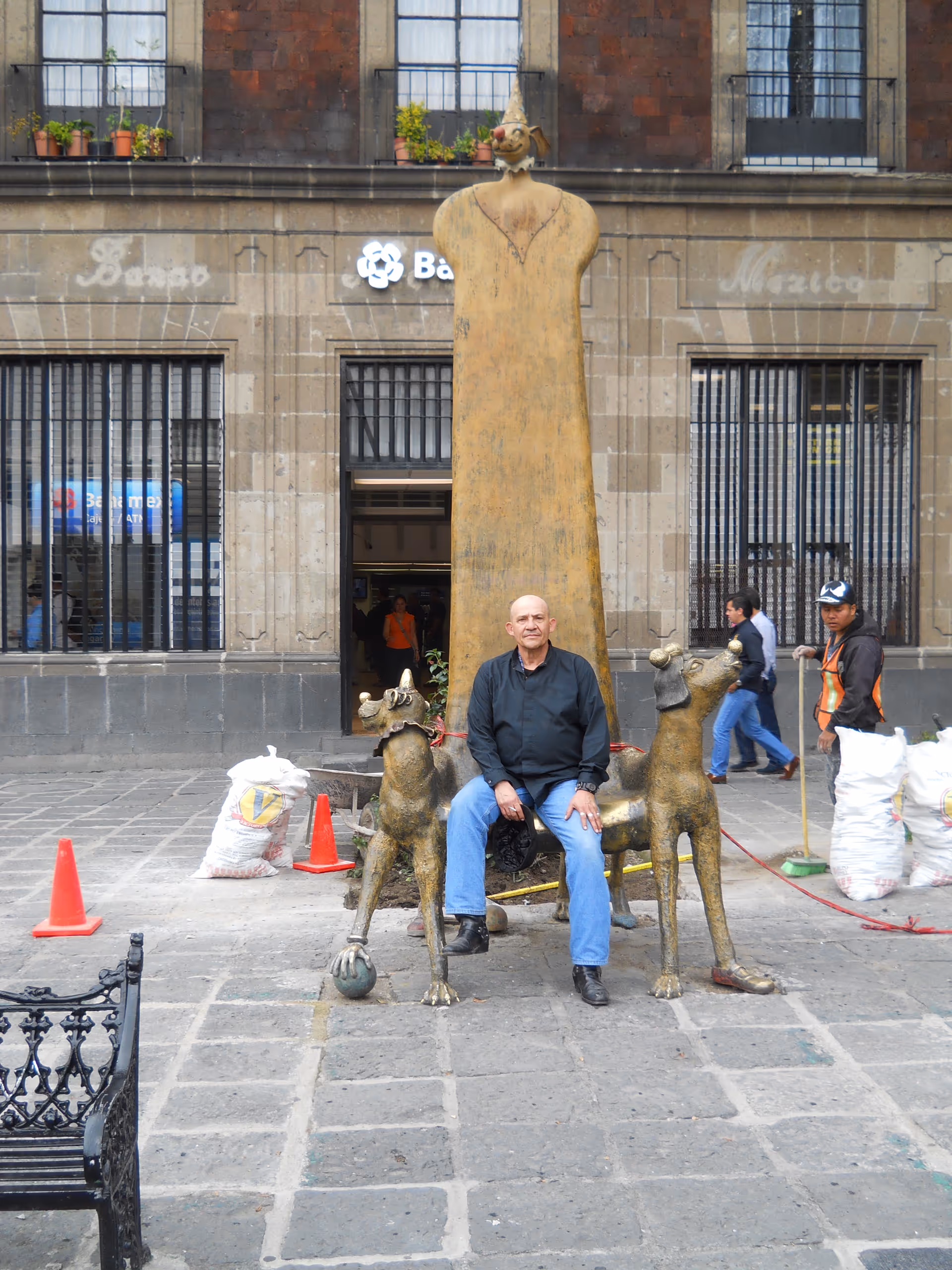 A man sits on an animal sculpture in a cobblestone plaza in front of a building.