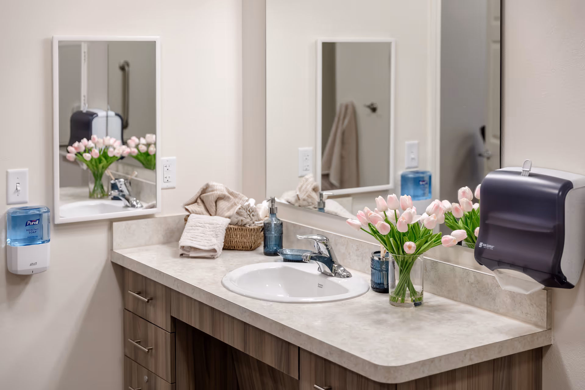 A clean bathroom countertop with a white sink, chrome faucet, and a large mirror above. On the countertop, there is a clear vase with pink tulips, a blue soap dispenser, a small blue dish, and a basket with folded beige towels. On the wall, there is a mounted Purell healthy soap dispenser and a paper towel dispenser.