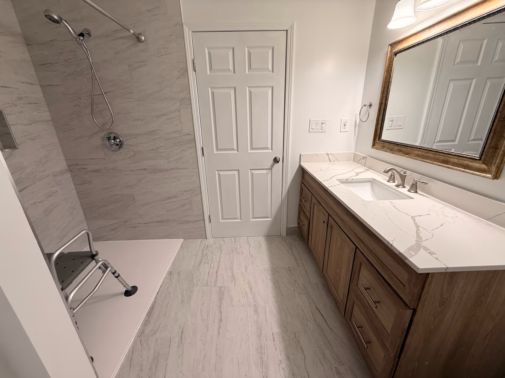Modern bathroom with marble walls, a wooden vanity, large mirror, and folding shower chair.