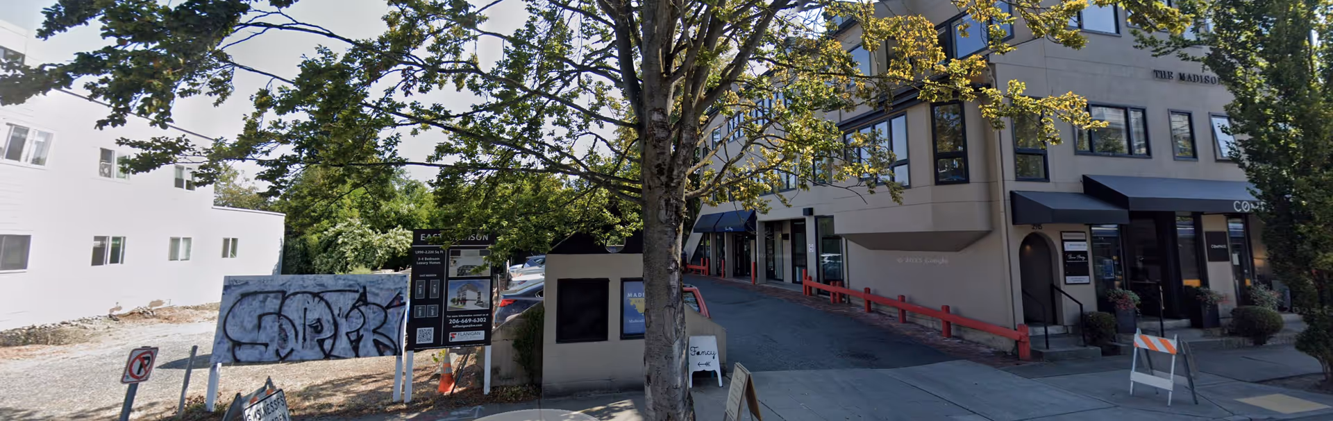 Exterior street view of a building with trees, storefronts, and construction signs.