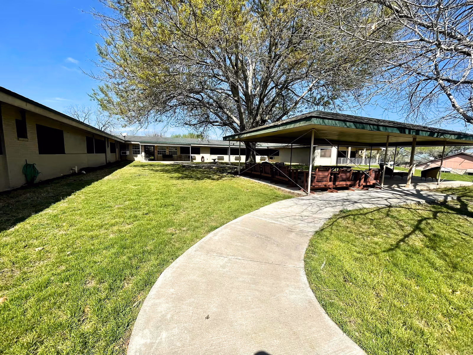 Outdoor area of Shiner Nursing & Rehab Center featuring a curved concrete walkway, green grass, a large tree with some leaves, and a covered seating area with wooden benches. The building surrounds the area on two sides under a clear blue sky.