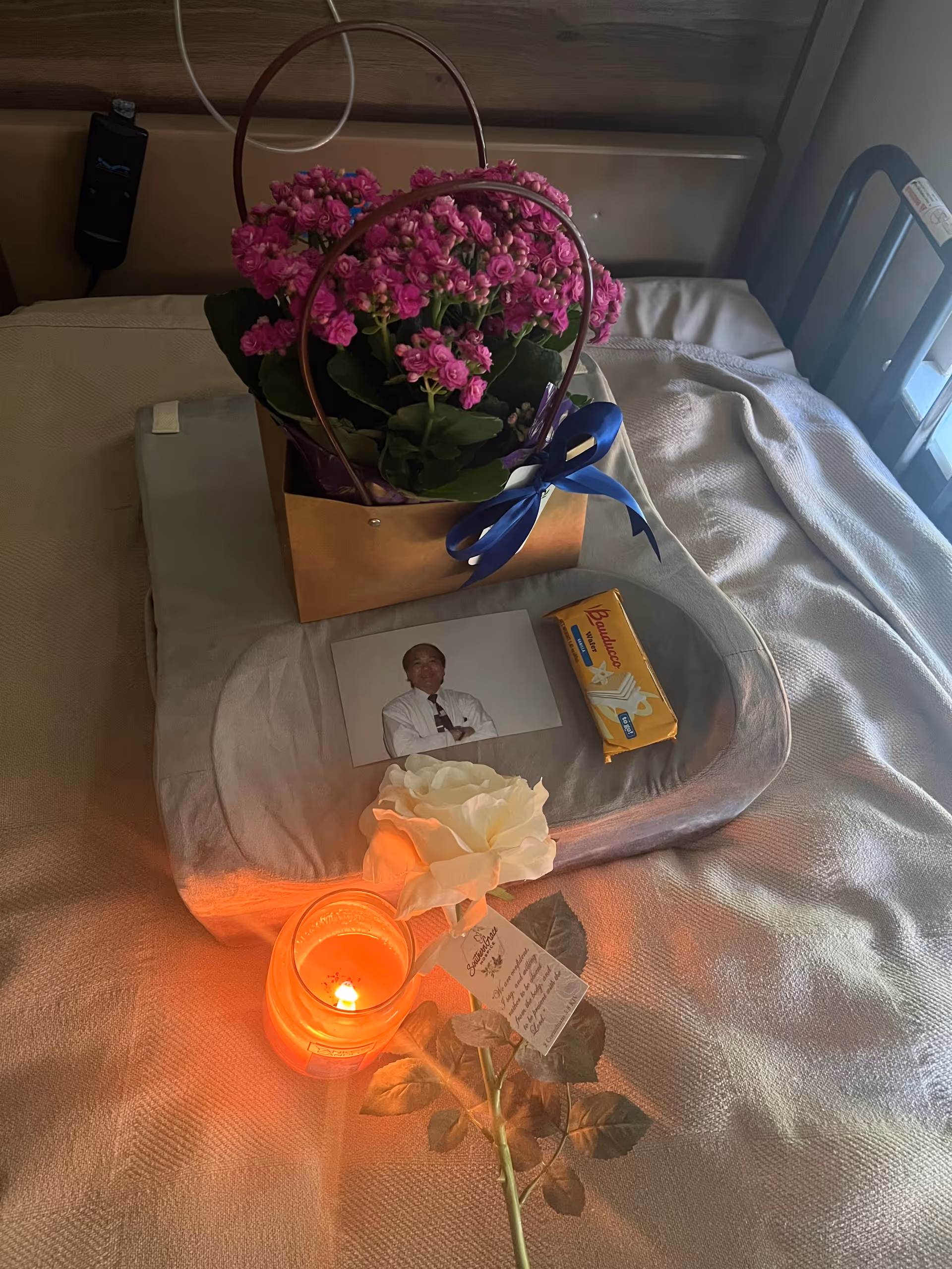 A hospice bed features pink bouquet, photo, candle, and card on the blanket.