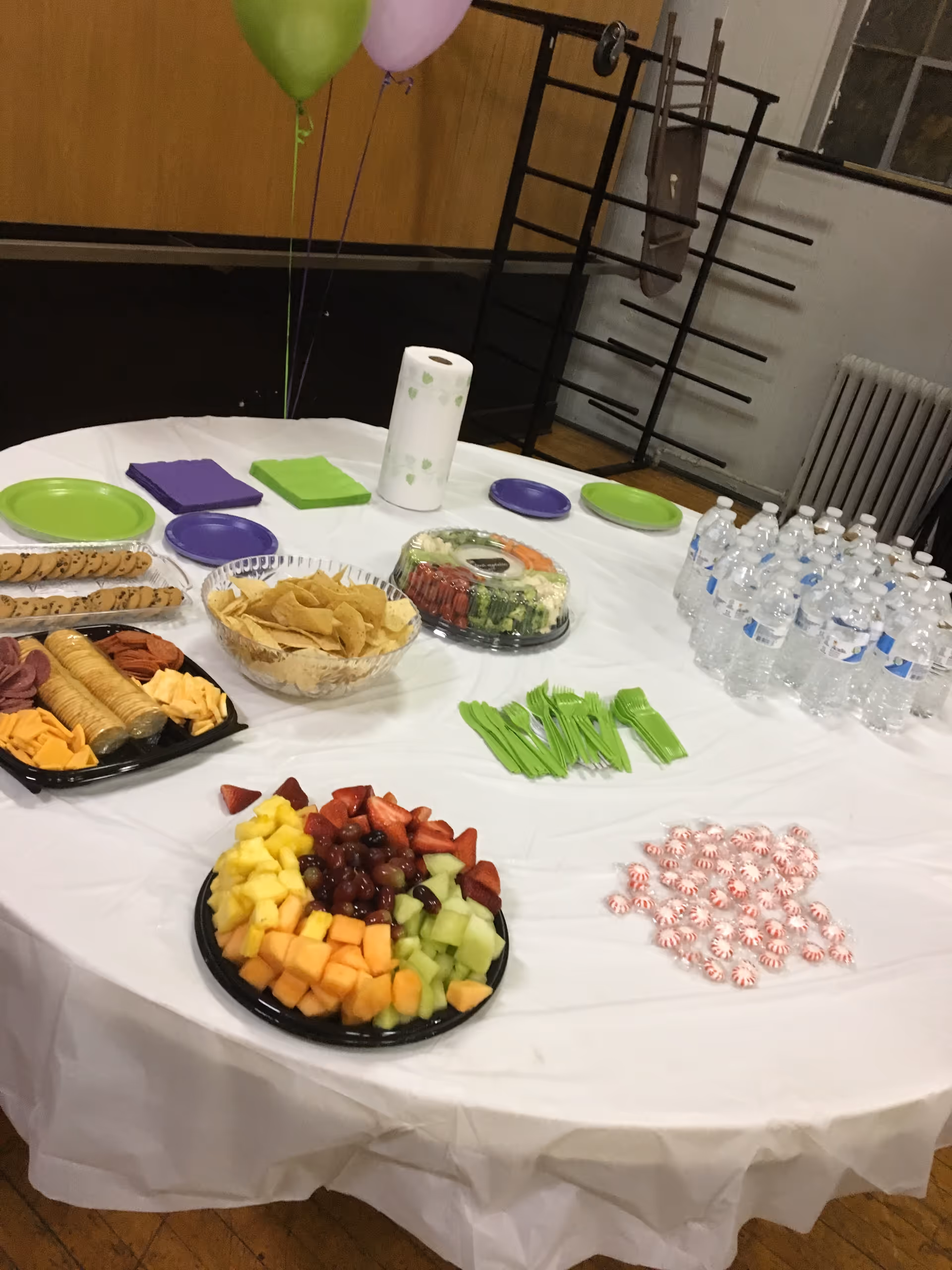 A round table covered with a white tablecloth holds fruit, snacks, water bottles, and balloons.