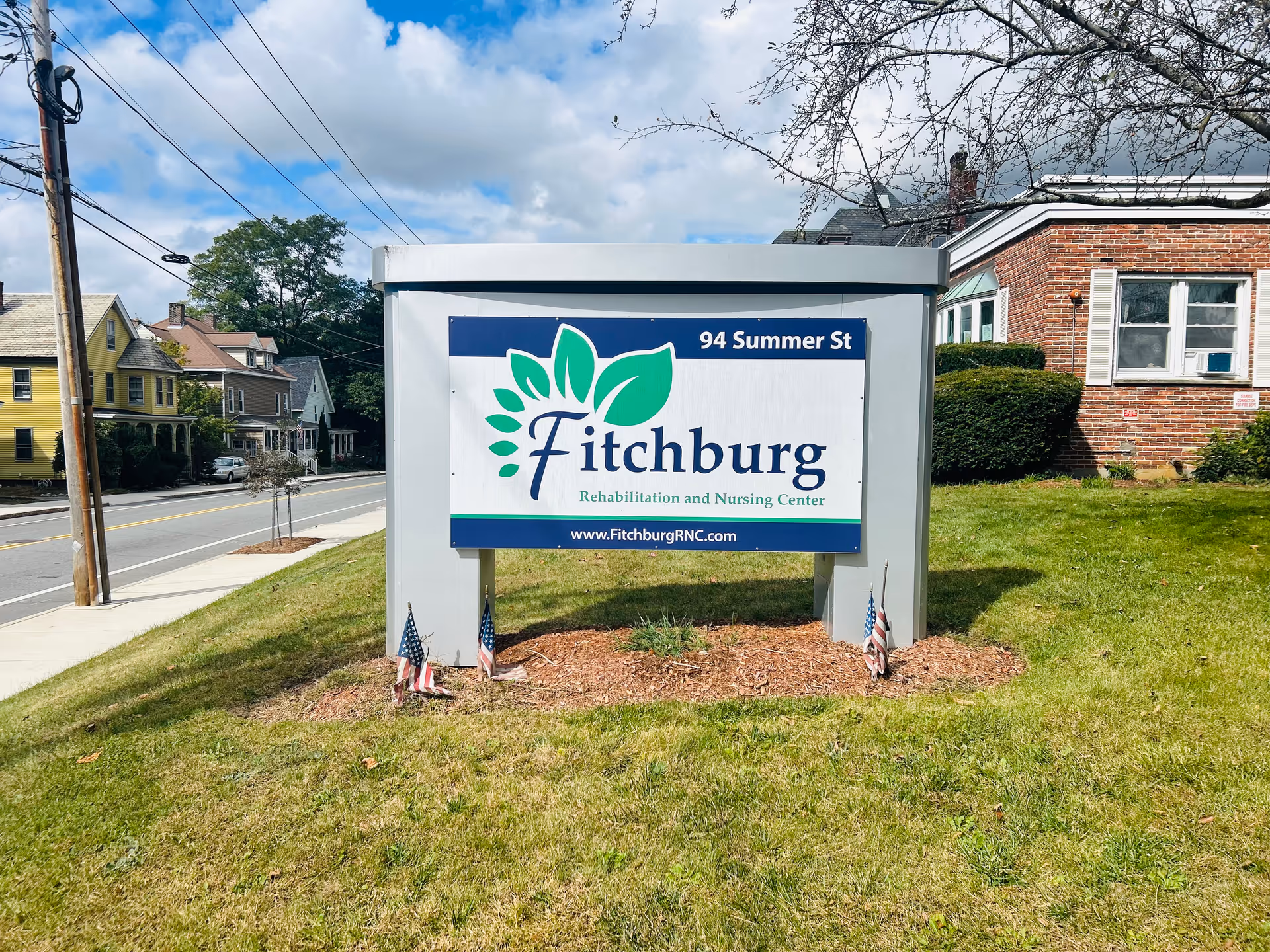 Outdoor sign for Fitchburg Rehabilitation and Nursing Center located at 94 Summer St, placed on a grassy area with small American flags around its base. The background shows a sidewalk, street, utility poles, houses, and a partly cloudy sky.
