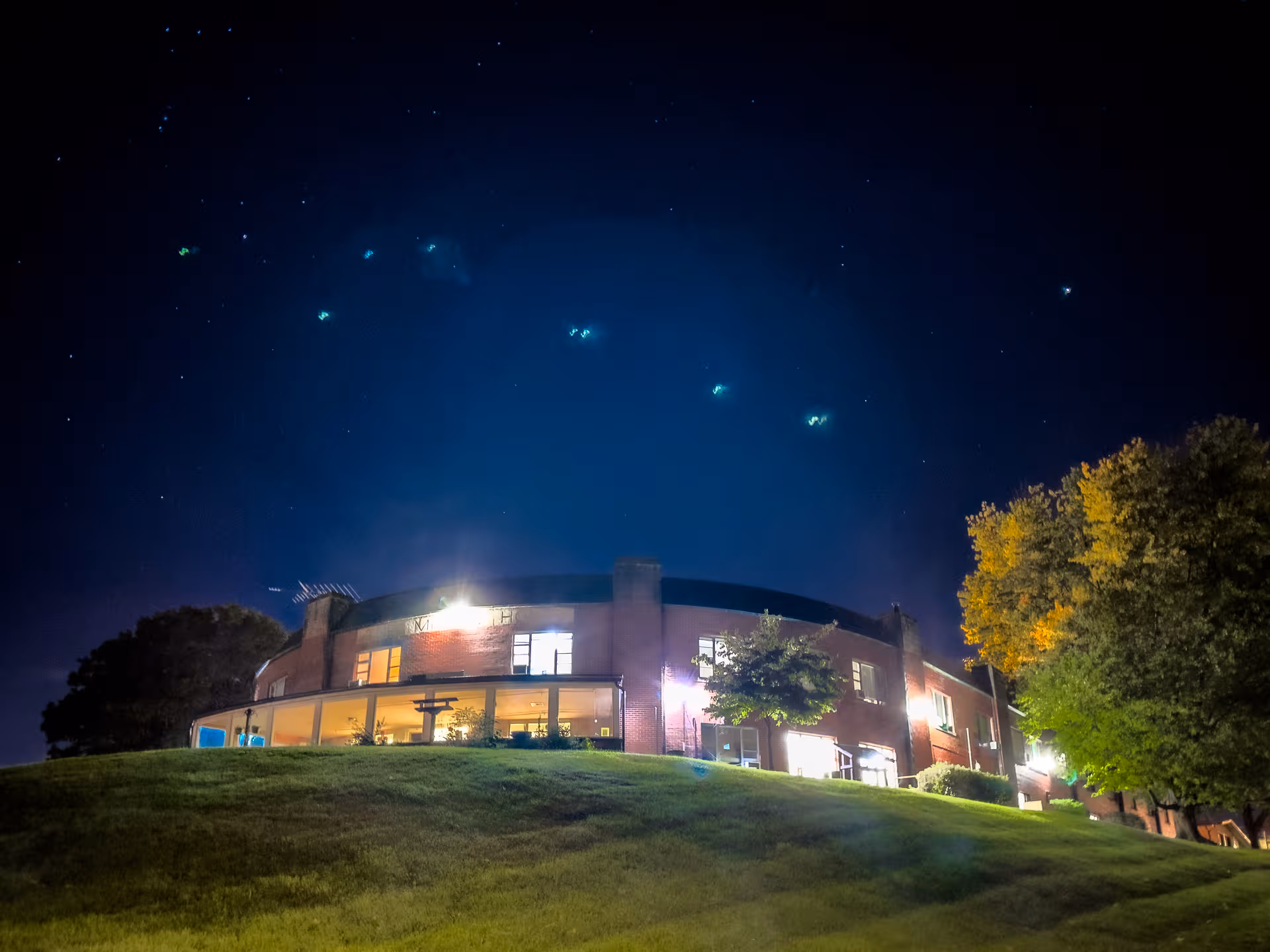 Night exterior of Milton Home: curved brick building on a grassy hill, lit windows.